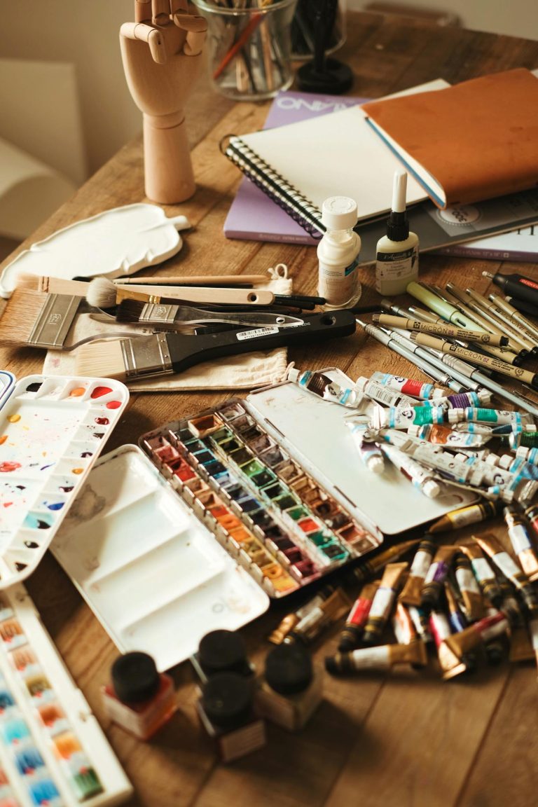 A collection of paints, brushes, and sketchbooks arranged on a wooden table in an art studio.